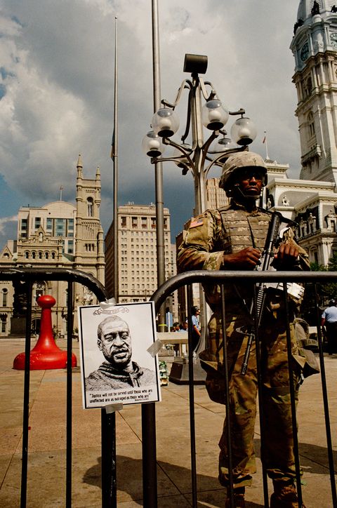 officer wearing camouflage and holding gun stands next to poster of george floyd