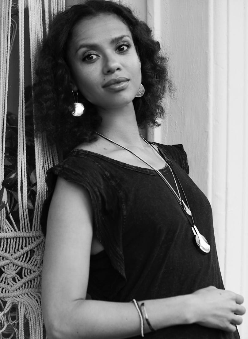 a person with curly hair wearing a dark shirt and jewelry leaning against a wall