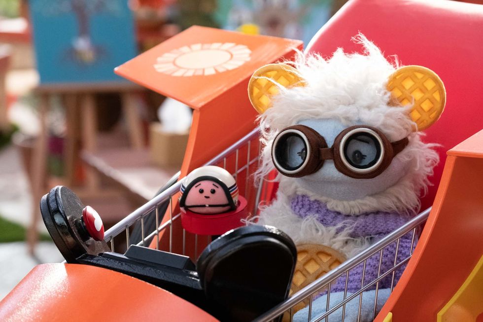 a furry white and blue puppet with frozen waffle ears, wearing protective flight goggles, is seated next to a pink, round mochi puppet, wearing a helmet they are inside a flying go kart smiling for the camera