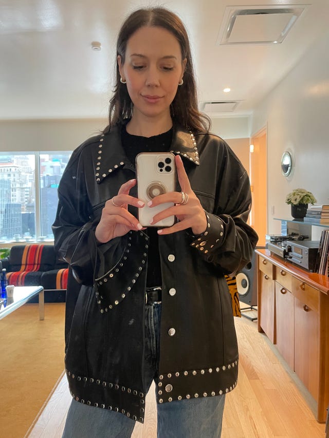 person holding a smartphone in front of a mirror wearing a studded black shirt jacket and jeans