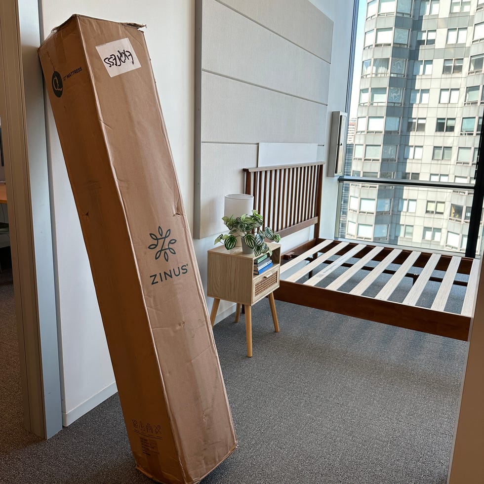 a box containing a zinus mattress is leaning against a wall in a room with a minimalistic bed frame and a nightstand