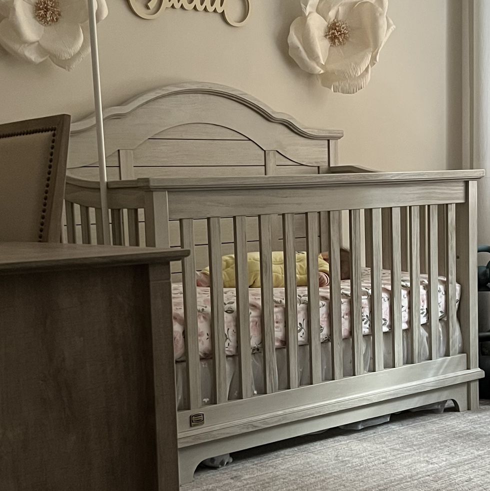 gray crib in a baby nursery with a baby sleeping inside under a wall decoration that says stella, part of good housekeeping's tips for safe crib use