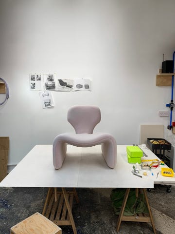 a minimalist chair with a soft curved design sits on a white table behind the chair several black and white sketches of various furniture pieces are pinned to a wall the workspace includes scattered tools such as scissors and measuring tape along with a green box and a green bag on the side the floor features a mix of textures indicating a creative environment
