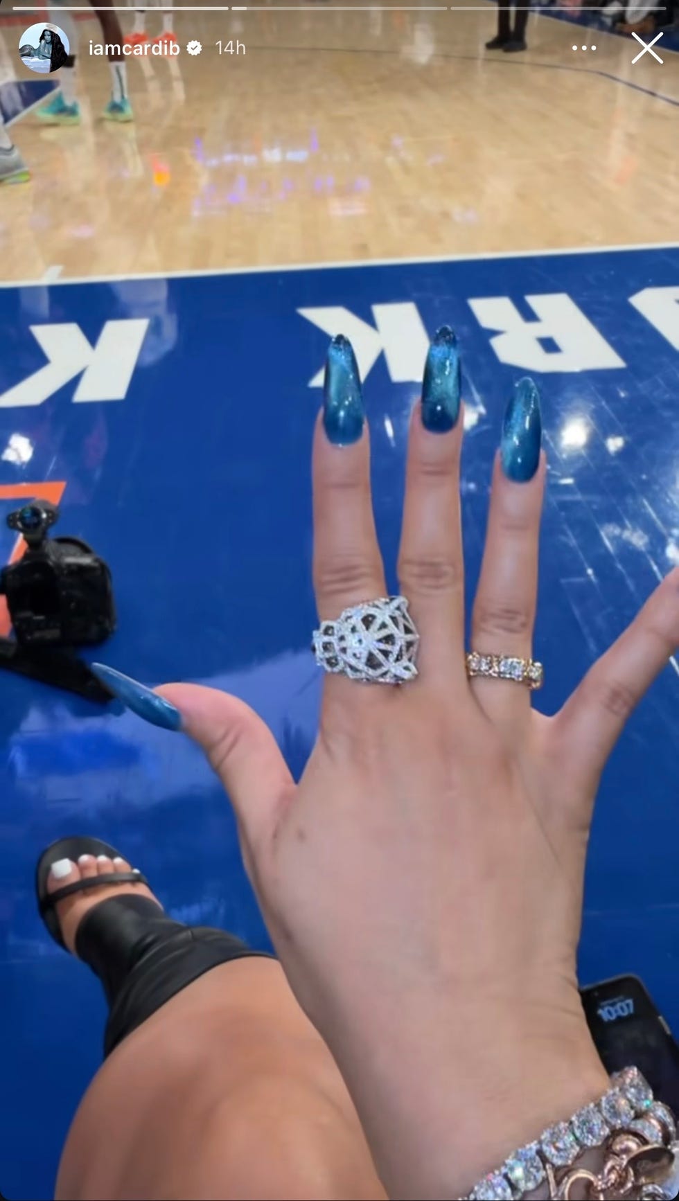 mão exibindo anéis com unhas brilhantes azuis em um ambiente esportivo