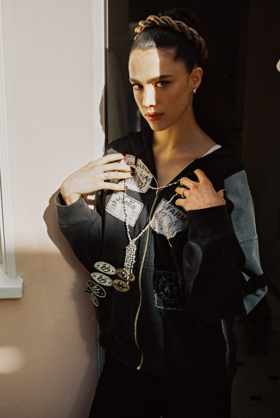 a person wearing a hooded sweatshirt with patches and jewelry posed with hands on shoulders in a softly lit room