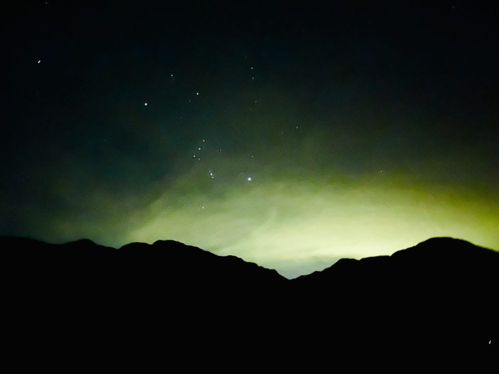 night sky with bright stars above dark mountain silhouettes and a faint glow on the horizon