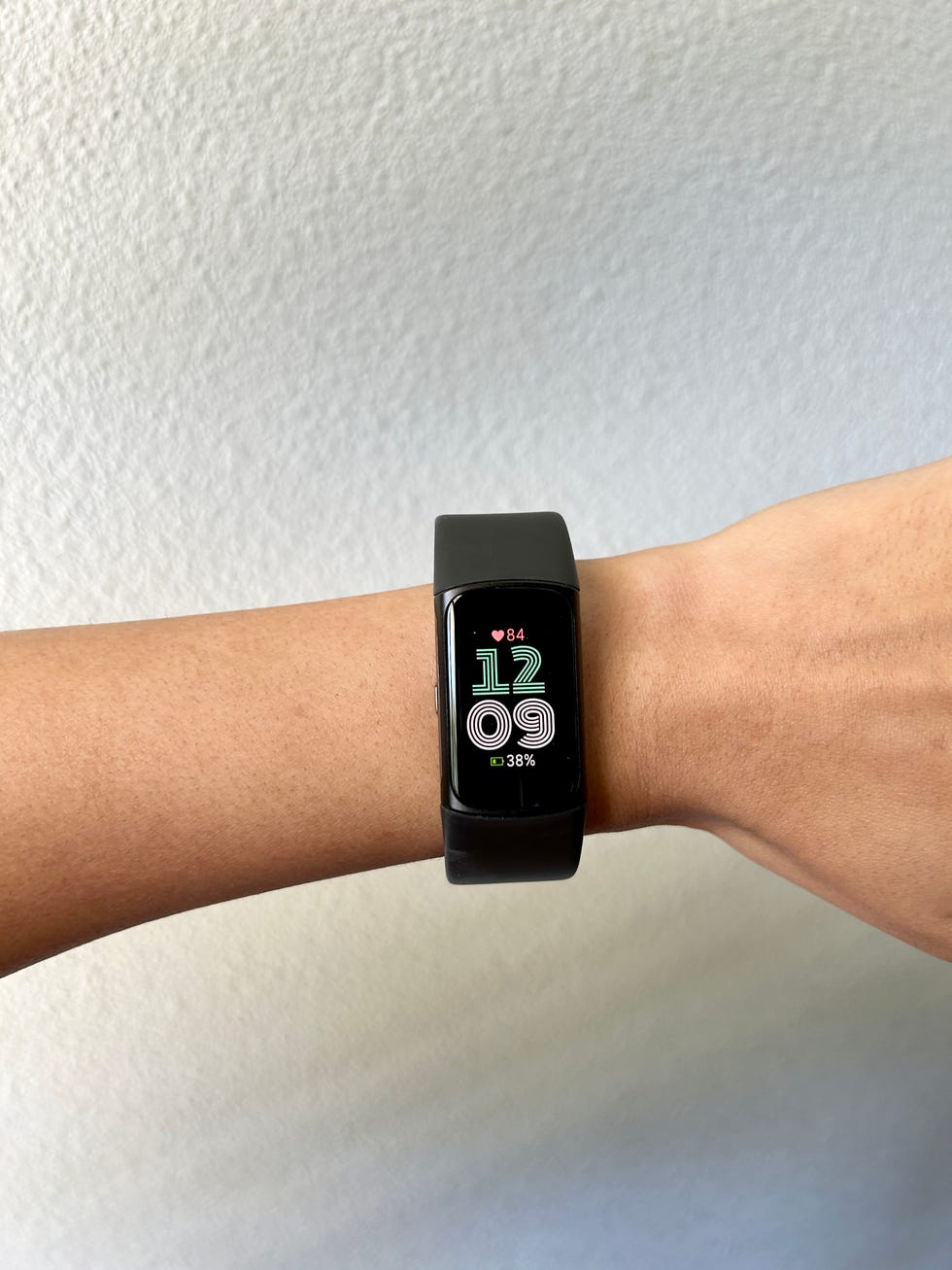 A black smartwatch is worn on the wrist. The display shows the time as 1209, heart rate of 84 beats per minute, and battery level of 38 percent. The background is a plain white wall.