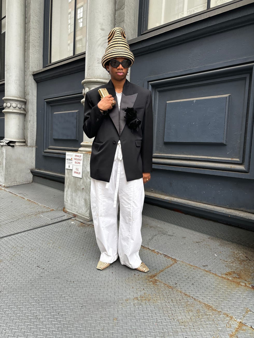 a person wearing a stylish outfit with a prominent hat stands outside a building