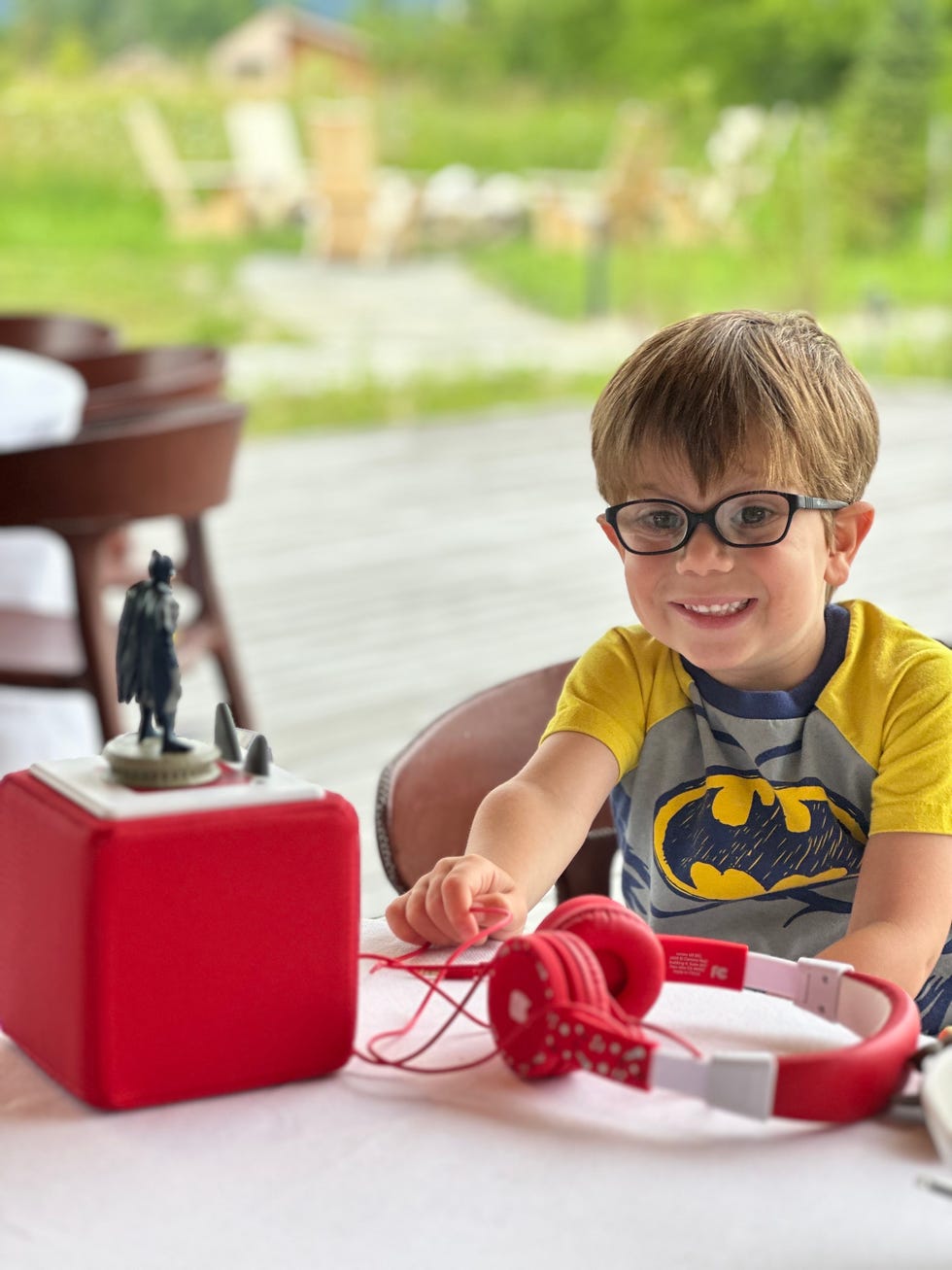 a young boy wearing glasses sits next to his batman tonie on a red toniebox, part of a good housekeeping review of the toniebox