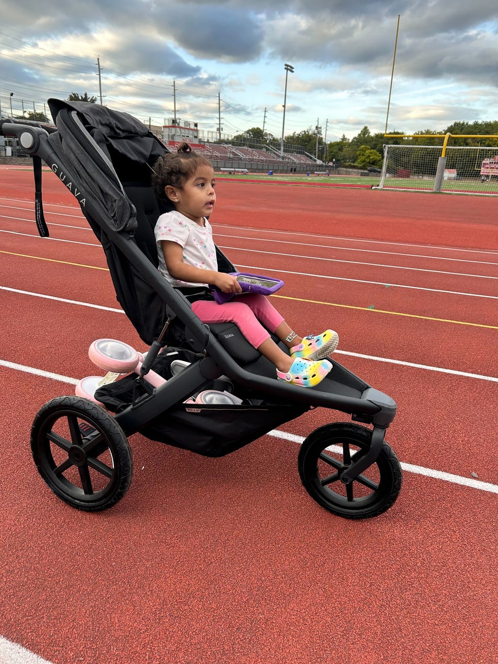 a girl in pink leggings and crocs sits in the guava roam jogging stroller to help her mom test it on a high school track