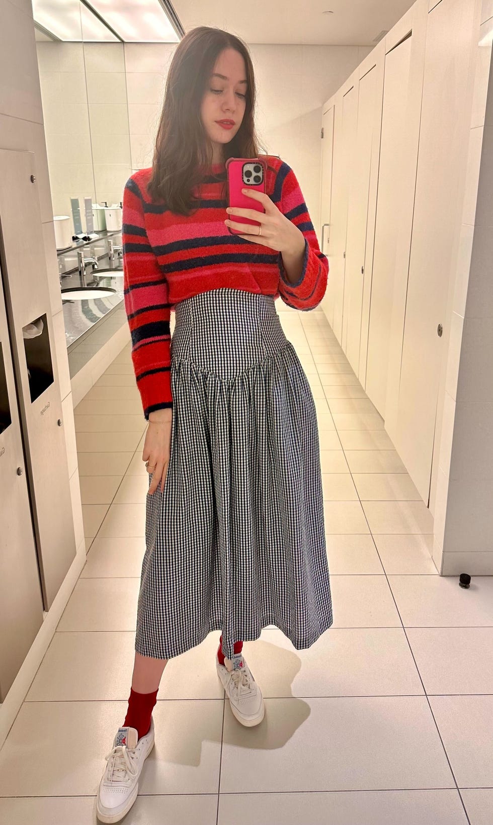 a gh analyst in a blue and white skirt and pink and blue striped sweater taking a selfie in the mirror