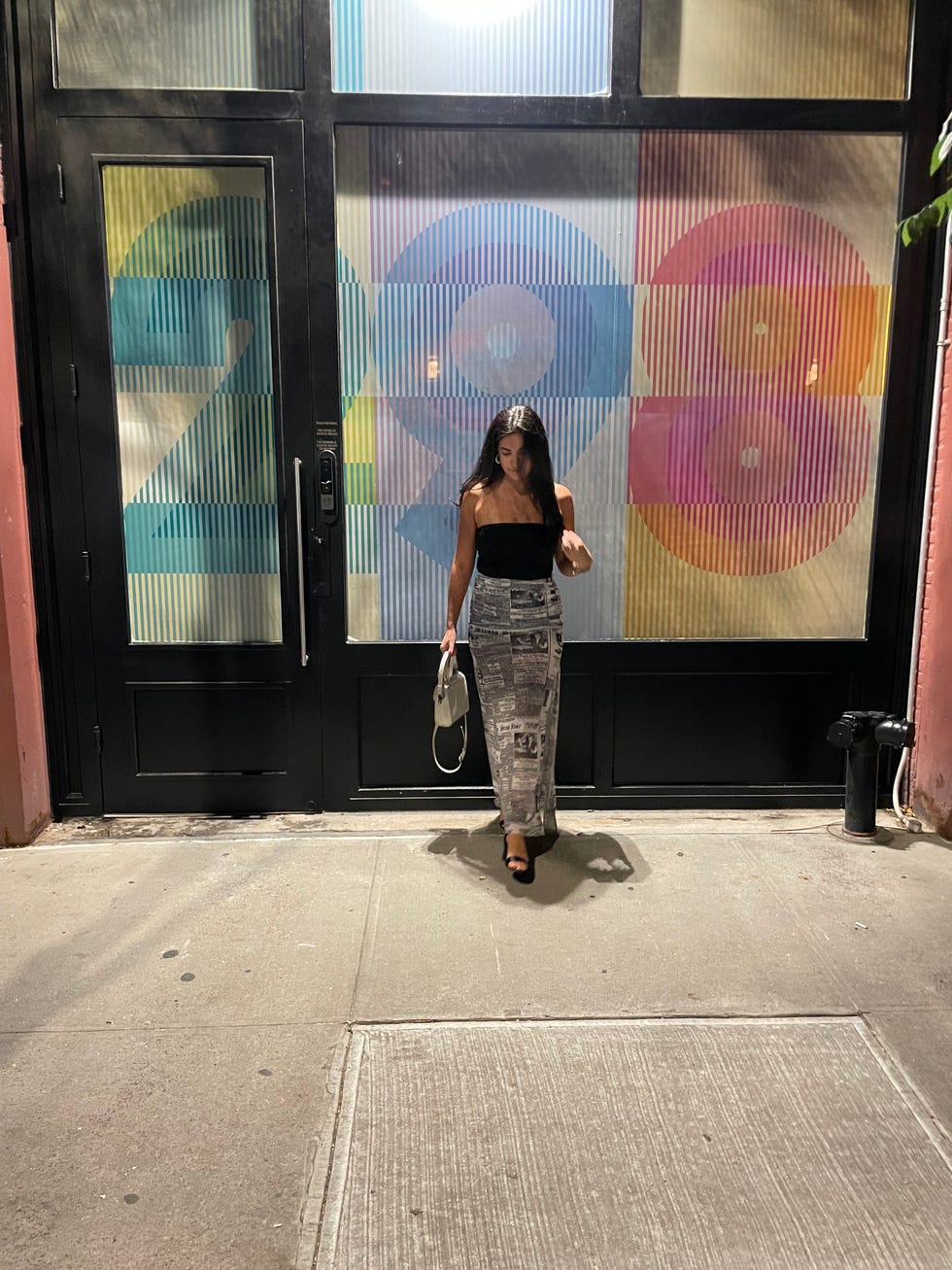 a person standing in front of a wall with a painting on it wearing a skirt from rent the runway with newspaper print, good housekeeping's rent the runway review