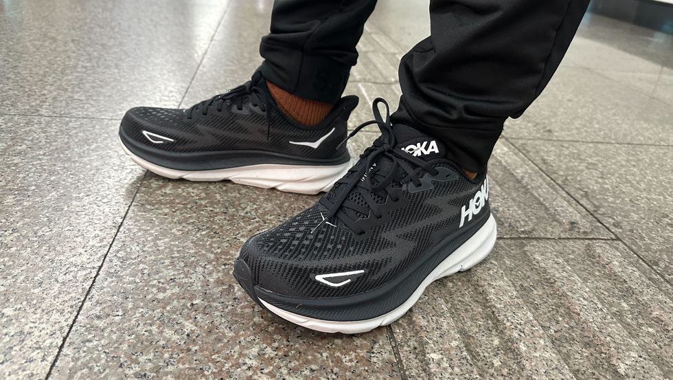 a tester wearing a pair of black and white hoka clifton 9 sneakers on grey tile as part of good housekeeping's testing for the best walking shoes for men