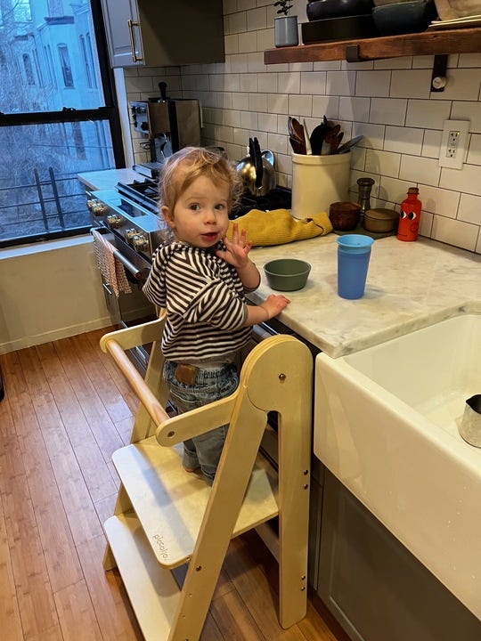 a toddler in a striped hoodie stands on the piccalio mini chef learning tower, part of a good housekeeping story on why a learning tower is worth it
