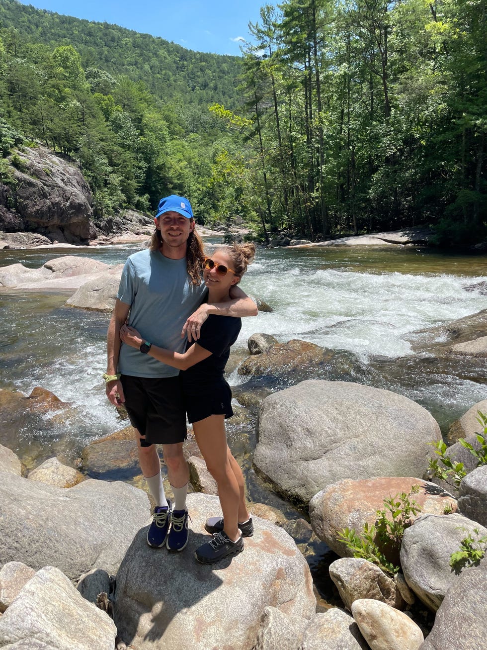 a couple is embracing while standing on large rocks along the edge of a flowing river in a lush green environment