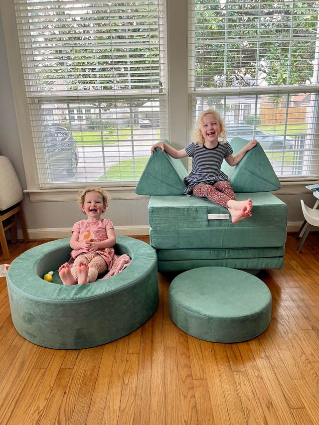two girls sit on a round nugget ottoman and a tall nugget play couch, both pieces of green play furniture that double as toys