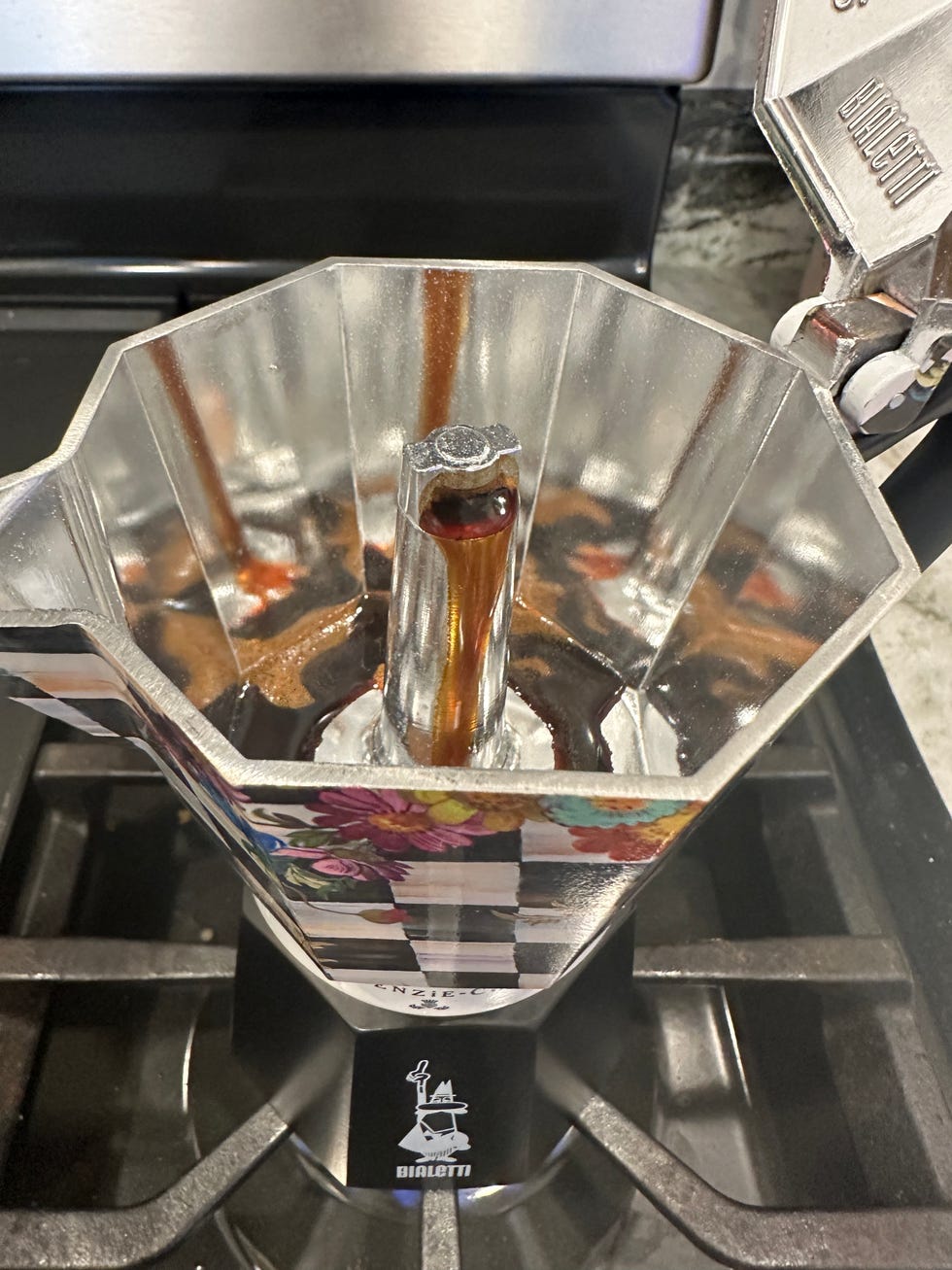 a moka pot sits on a stovetop with brewed coffee dripping from the spout into the upper chamber the pot features a decorative design with flowers and stripes, and the liquid is dark and rich in color the surrounding area shows elements of a kitchen countertop