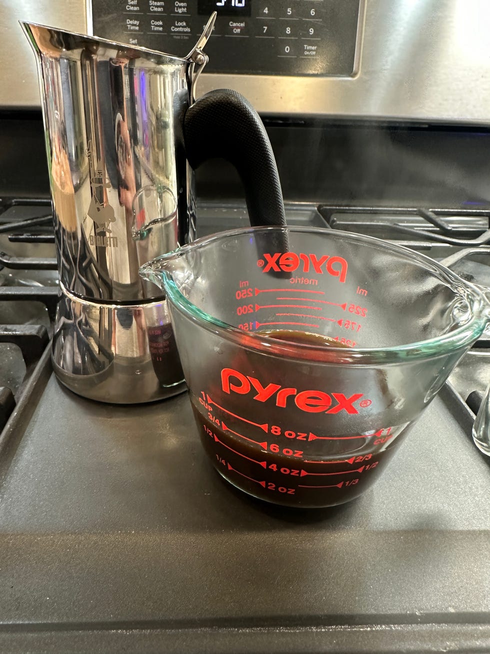 a stainless steel stovetop espresso maker is positioned next to a glass measuring cup filled with freshly brewed coffee steam is rising from the coffee, indicating it is hot the measuring cup has red markings for measurement, showing the volume of coffee inside the background features a black stove top and a digital oven display, indicating time and settings