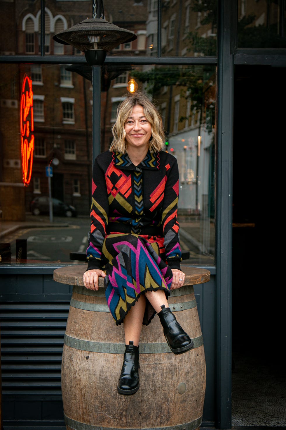 Person sitting on a wooden barrel in front of a rustic storefront with neon light