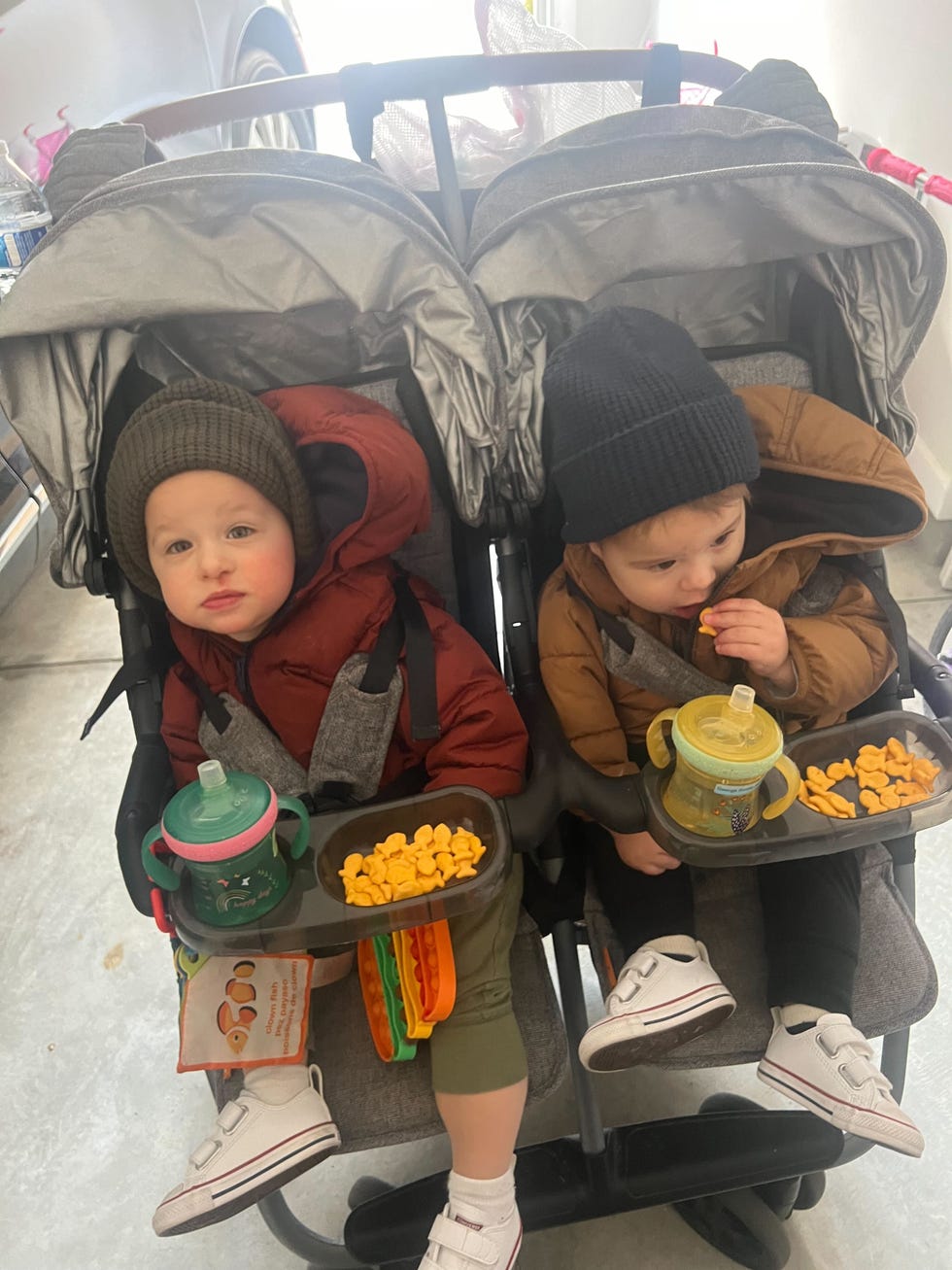 twin babies wearing hats enjoy their snacks and sippy cups in a zoe twin stroller, part of a good housekeeping story on the best double strollers