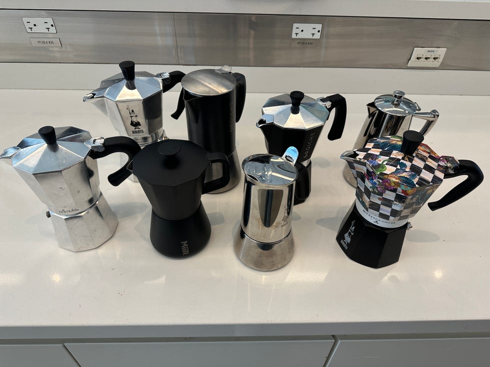a variety of stovetop espresso makers are displayed on a modern kitchen countertop the collection includes several aluminum and black models in different designs and sizes some have shiny metallic finishes while others feature matte surfaces one unique item has a colorful checkerboard pattern, adding a distinct visual element to the group the countertop is smooth and reflects light, enhancing the overall contemporary kitchen setting