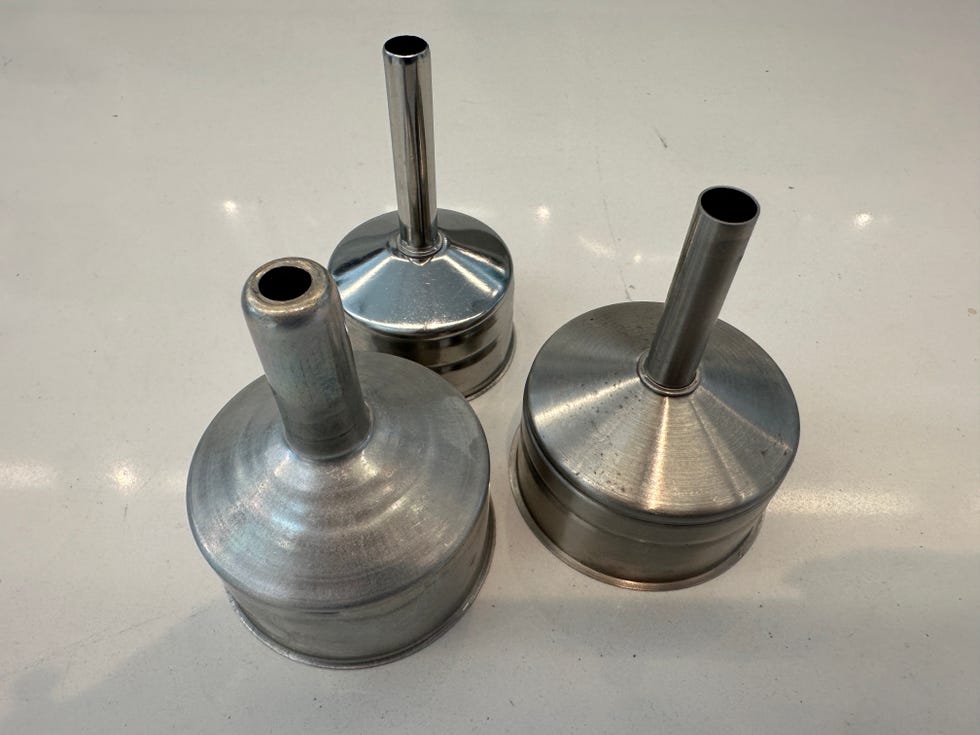 three metallic funnels are arranged closely together each funnel has a conical shape leading to a cylindrical opening at the top the surfaces appear smooth and shiny, reflecting light, and they differ slightly in size and height they are positioned on a light colored, flat surface