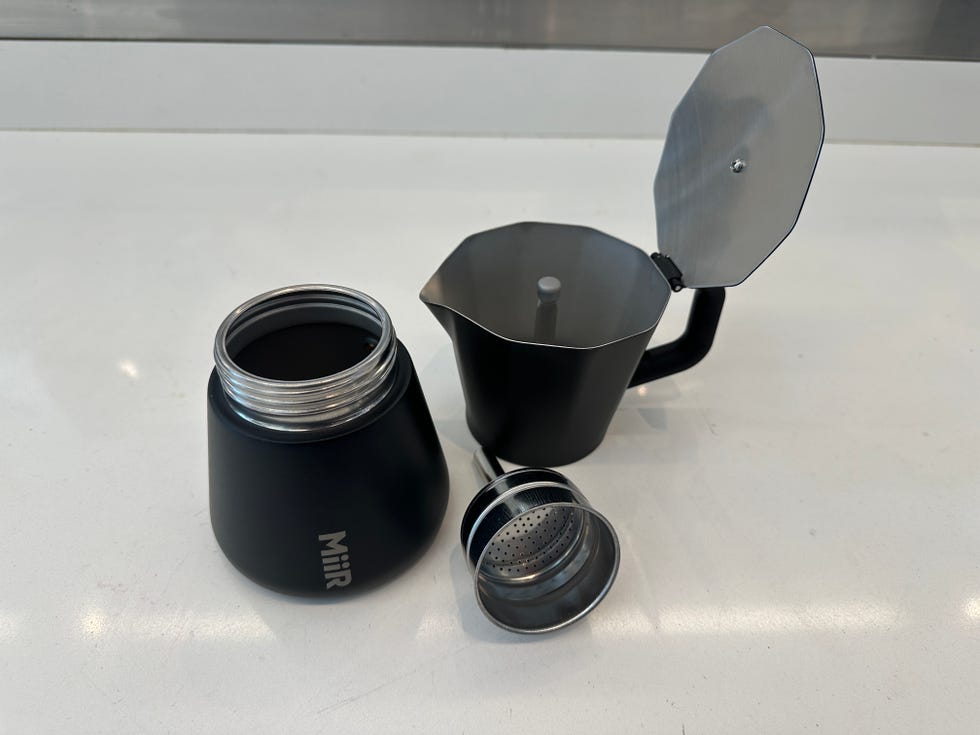 a thermal coffee container with a black matte finish is partially disassembled, revealing its metal threaded opening next to it is a black moka pot with a metal handle and a slightly open lid, which showcases its brewing chamber a shiny metal filter basket sits nearby, used for holding coffee grounds during the brewing process