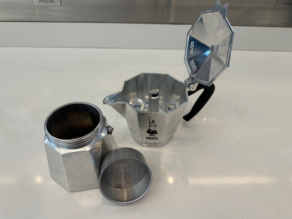 an aluminum moka pot featuring a hexagonal base and a pouring spout, with the upper chamber detached the lower section shows a small opening with a screw, and the filter disk is lying nearby the surface beneath is smooth and light colored, providing a clean backdrop for the coffee maker components