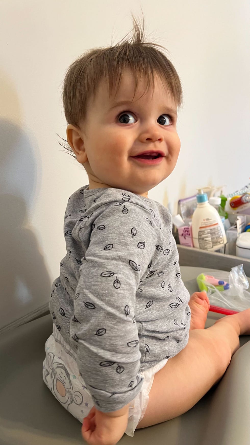 a toddler wearing a gray shirt with leaf patterns seated on a changing table in his diaper