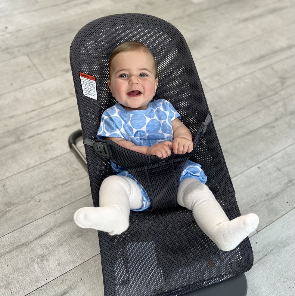 a baby in a babybjorn bouncer seat made of black mesh on a wood floor, for the good housekeeping review of best bouncer seats