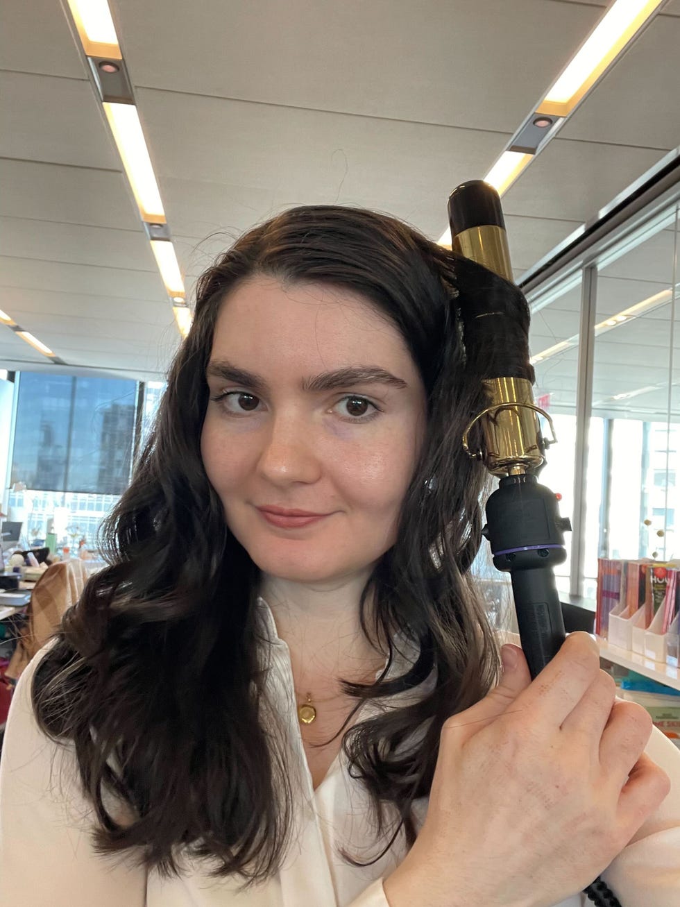 a person is holding a curling iron with a gold barrel, positioned near their long, wavy dark hair