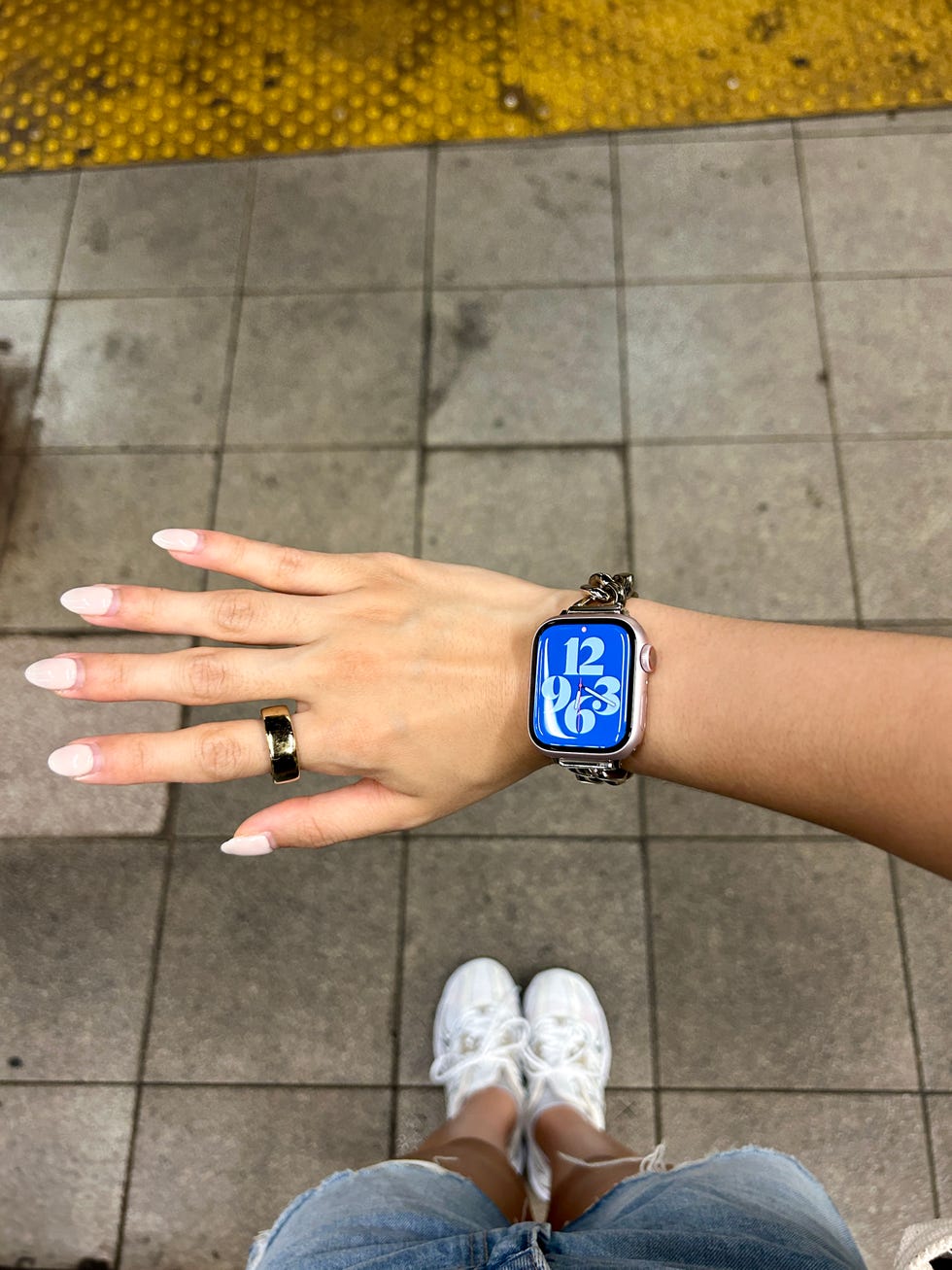 A person's hand is prominently displayed, along with a bright blue background and a smartwatch with large white numbers. A person's legs are partially visible in denim shorts and white sneakers.