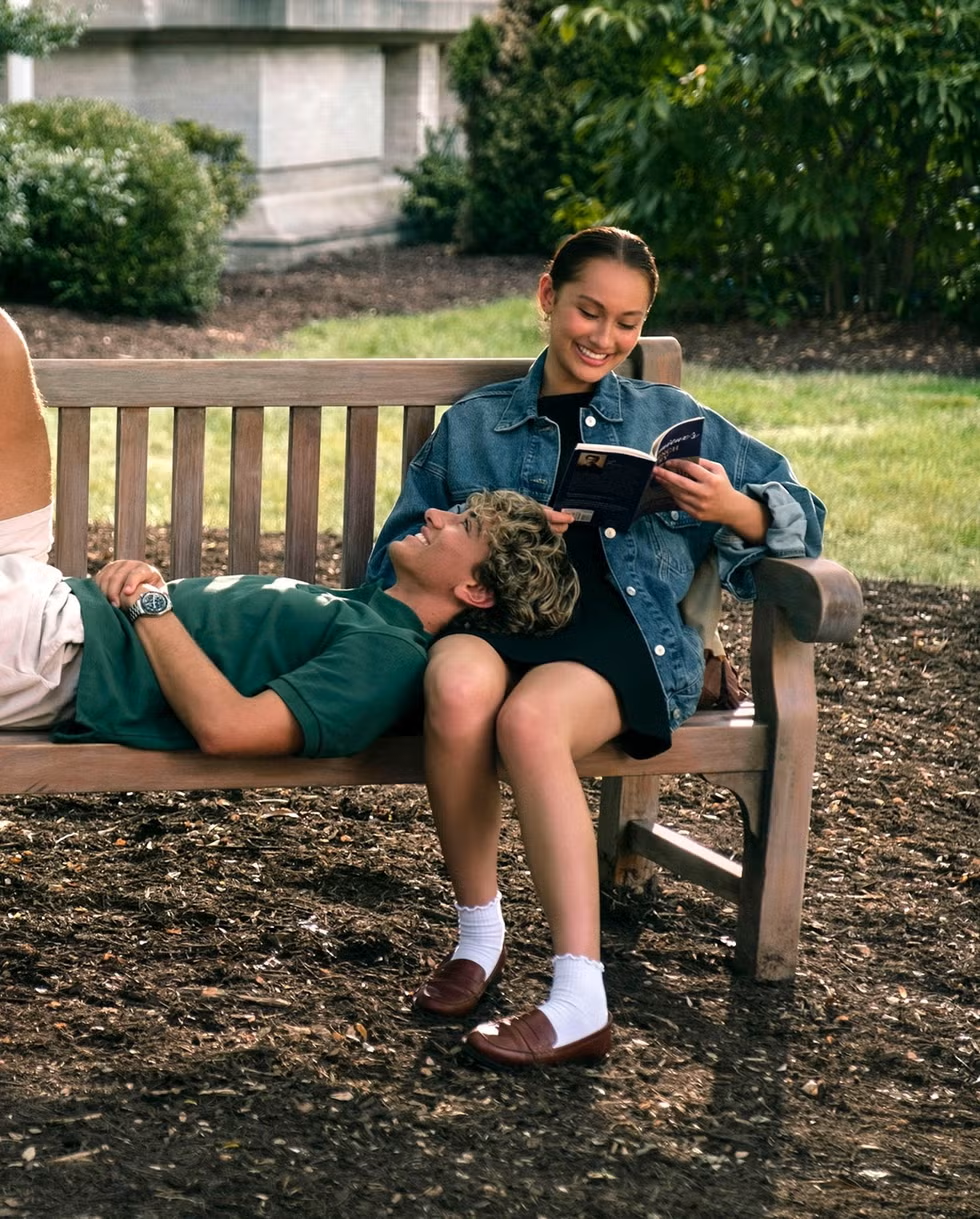 a girl reading a book on a bench with a man's head in her lap