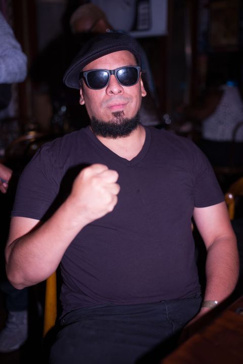 immortal technique wears a black t shirt, dark sunglasses, and hat he raises a fist and smiles slightly