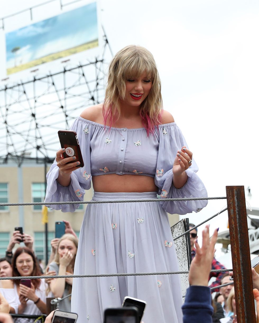 a woman smiles as she looks down at a crowd of people from behind a roped off platform, she holds a cell phone in one hand and has pink dyed tips in her hair