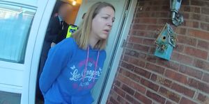 lucy letby, wearing a blue sweatshirt, is escorted in handcuffs from a brick house by a police officer, whose face is obscured