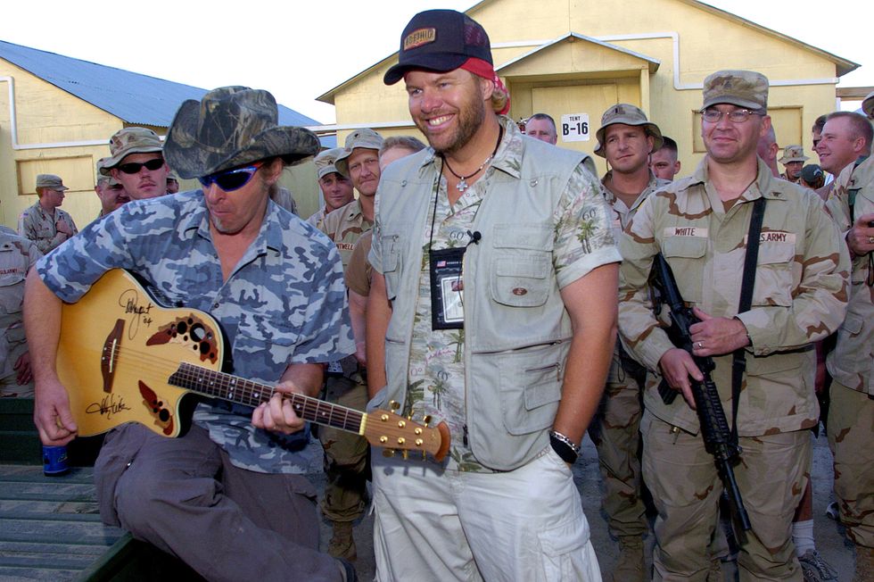ted nugent strums a guitar as he sits next to a standing toby keith who smiles with his hands in his pockets, behind them is a group of military personnel and two buildings