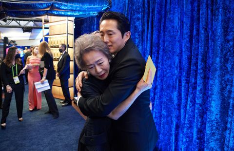 steven yeun giving youn yuhjung a hug in front o a blue curtain