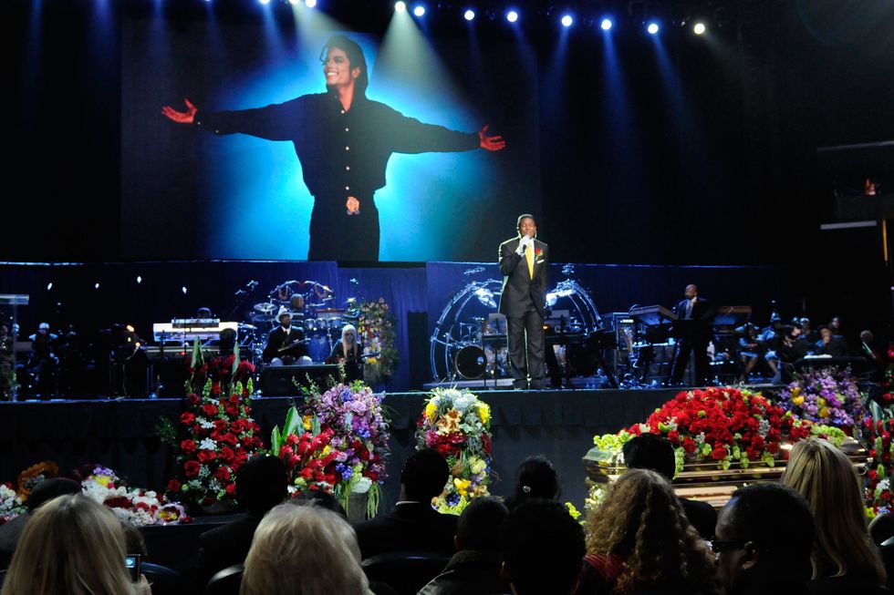 jermaine jackson, wearing a black suit and yellow tie, sings into a microphone on a stage, with a large photo of michael jackson projected onto the wall behind him, and several flowers and a coffin in front of him