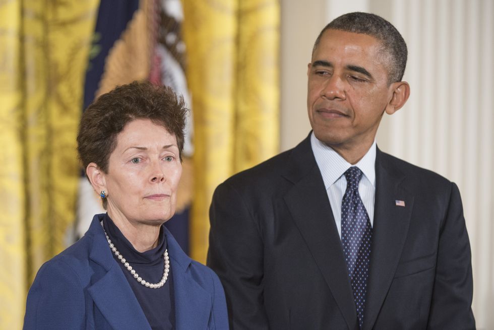 a woman and a man stand next to each other, both wear suits, she looks ahead as he looks to the left at her