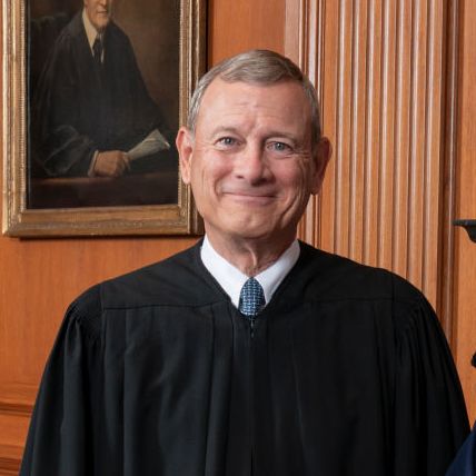 supreme court chief justice john roberts smiles at the camera, he is wearing his black justice robes with a blue tie, behind him is a wood paneled wall with a partial portrait in view
