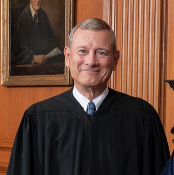 supreme court chief justice john roberts smiles at the camera, he is wearing his black justice robes with a blue tie, behind him is a wood paneled wall with a partial portrait in view