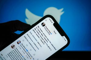 an image of a shadowy hand holding a phone, which has tweets by stephen king and elon musk on it, with a blue wall backdrop with the twitter logo on it