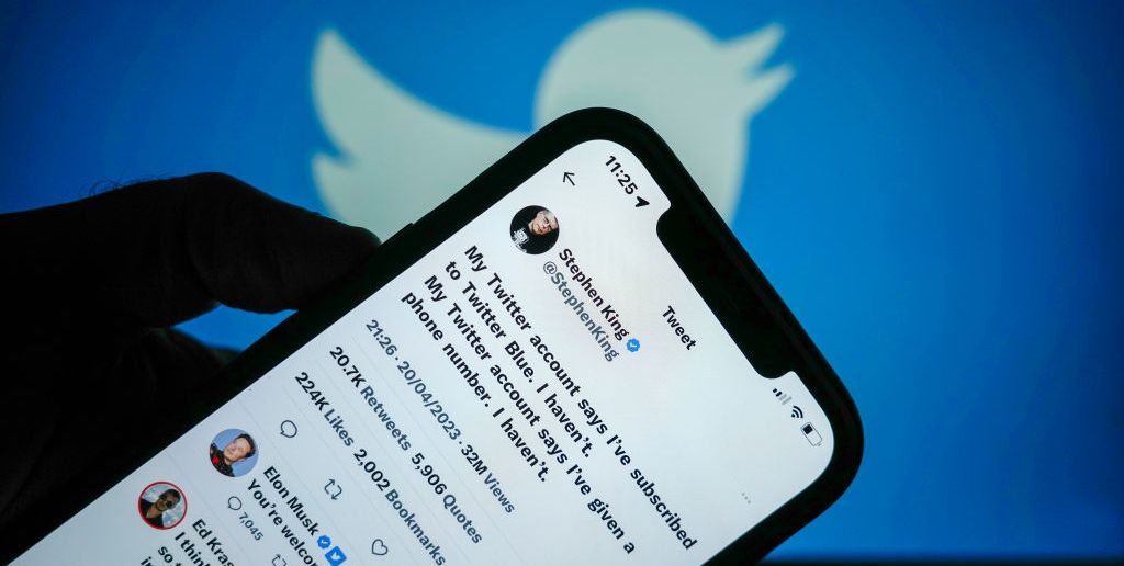 an image of a shadowy hand holding a phone, which has tweets by stephen king and elon musk on it, with a blue wall backdrop with the twitter logo on it