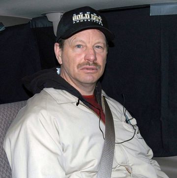 gary ridgeway looks at the camera while buckled into a car seat, he wears a dark ball cap, tan jacket and red shirt