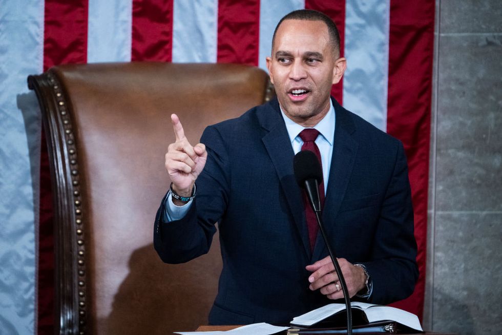 hakeem jeffries stands at a lectern and speaks into a microphone while pointing one finger upward, he wears a navy suit jacket and red tie, behind him is a large brown leather chair and part of a large american flag