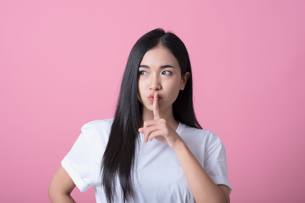 index finger on lips, silence gesture, shhh quiet, asks for voicelessness forefinger at the mouth young attractive woman in a light t shirt on pink background