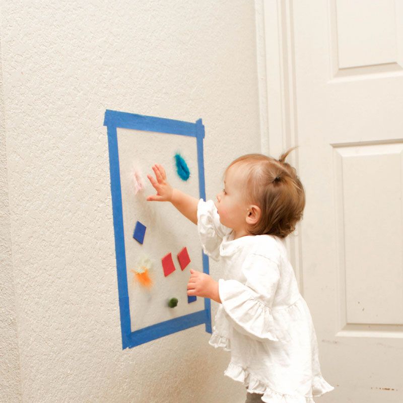 a toddler sticks shapes to a sheet of contact paper attached to a wall with the sticky side out