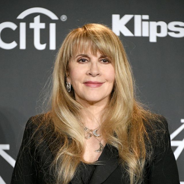 stevie nicks smiles and looks just past the camera while standing in front of a dark gray backdrop, she wears all black, several silver and gold necklaces, and large dangling earrings, her long blond hair is down