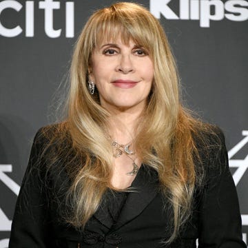 Stevie Nicks at the 2019 Rock & Roll Hall of Fame Induction Ceremony stevie nicks smiles and looks just past the camera while standing in front of a dark gray backdrop, she wears all black, several silver and gold necklaces, and large dangling earrings, her long blond hair is down